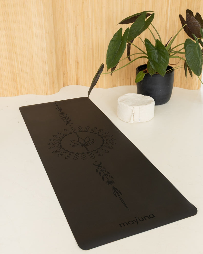 Mayuna Yogamatte Lotus in Schwarz mit Mandala-Gravur, ausgelegt neben einer Monstera-Pflanze