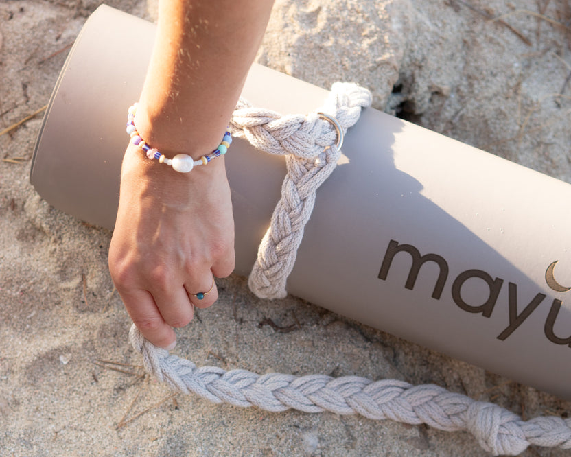 Hand hält den Mayuna Makramee-Tragegurt an einer zusammengerollten Yogamatte im Sand
